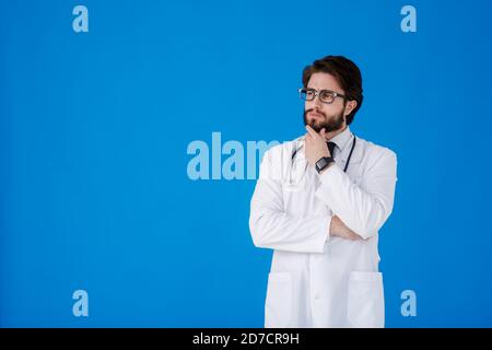Ein junger bärtiger Arzt auf blauem Hintergrund in einem weißen Mantel schaut auf die Kamera. COVID-19. Selbstisolierung. Bleib zu Hause Konzept. Quarantäne. Banner Stockfoto
