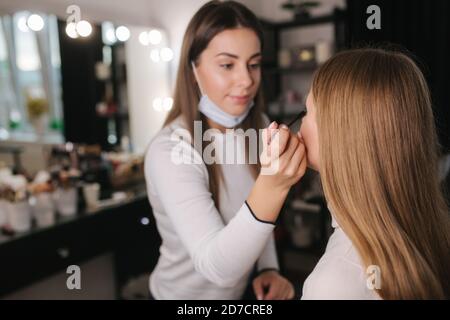 Weibliche Make-up-Künstlerin arbeitet im Schönheitssalon während der Quarantäne. Make-up Künstler in medizinische Maske auf Kinn. Schöne blonde Haare Modell Stockfoto