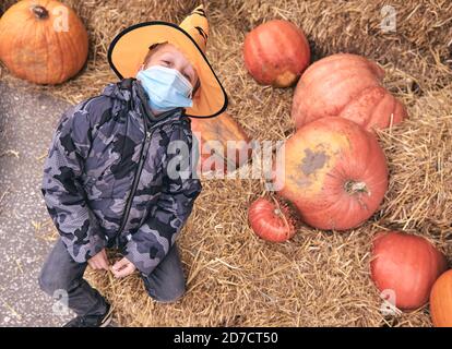 Junge im Halloween Kostüm mit Kürbissen auf dem Bauernmarkt steht auf Heu. Furchterregende Dekorationen. Kinder Trick or Treat. Das Tragen einer schützenden Gesichtsmaske an Halloween Stockfoto