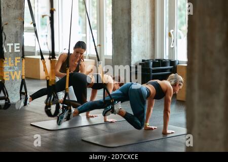 Körperliche Stärke entwickeln. Zwei junge sportliche Mädchen trainieren mit trx Fitness-Gurten mit weiblichem Personal Trainer im Fitnessstudio. Sport, Training, Wellness und gesunder Lebensstil Stockfoto