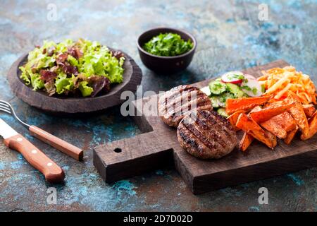 Gegrillte Burger-Cutlets mit gebackenen Süßkartoffeln, Gemüse und Salat auf Holzschneidebrett serviert, selektiver Fokus Stockfoto