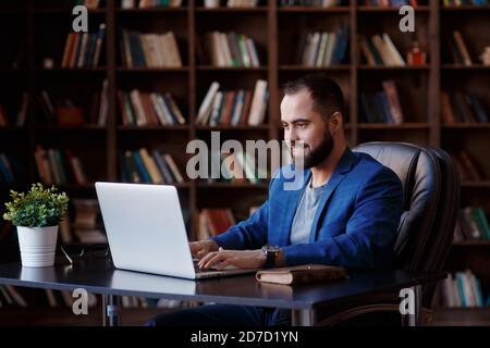 Ein junger Geschäftsmann arbeitet in einem luxuriösen Büro in der Bibliothek an einem Laptop. Ein bärtiger, fröhlicher Mann in einem blauen Anzug an einem Schreibtisch tippt auf einer Tastatur Stockfoto