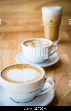 Zwei Tassen heißen Cappuccino-Kaffee mit Herzkaffee rt Oben mit Milch Eiskaffee im Hintergrund Stockfoto