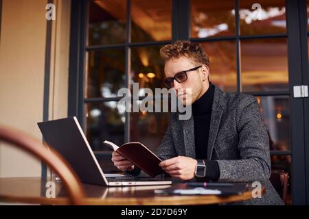 Elegant junge Kerl in formelle Kleidung sitzt im Café mit Sein Laptop und liest das Buch Stockfoto