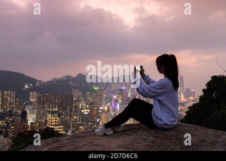 Silhouette eines Mädchens, das am Felsen sitzt und Fotos von der nächtlichen Ansicht der Stadt Skyline von Hong Kong Island (südlich von Victoria Harbour) vom Braemar Hügel macht, Stockfoto