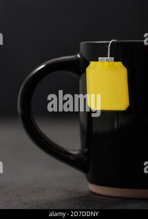Teebeutel mit gelbem leeren Tag von schwarzem Tee in schwarzer Keramik-Tasse auf dunklem Hintergrund. Stockfoto