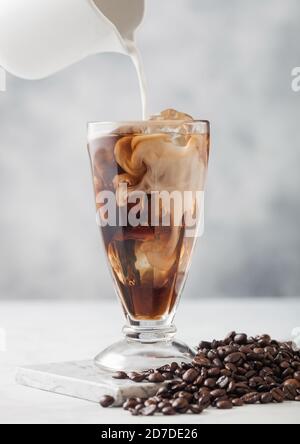 Gießen Sie Milch in Milchshake klassischen Glas mit eisgekühltem schwarzen Kaffee und frischen rohen Bohnen auf hellem Tischhintergrund. Stockfoto
