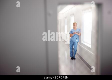 Weibliche Doctor Trägt Scrubs Im Krankenhaus Korridor Mit Digital Tablet Blick Durch Fenster In Der Tür Stockfoto