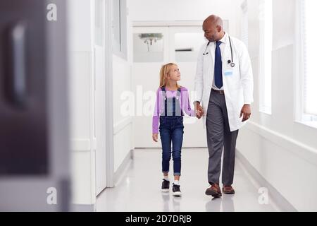 Männlich Pädiatrie Arzt Zu Fuß Entlang Krankenhaus Korridor Halten Hände Mit Junge Patientin Stockfoto