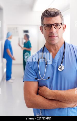 Portrait Von Reife Männliche Arzt Tragen Scrubs Stehen In Beschäftigt Krankenhauskorridor Stockfoto