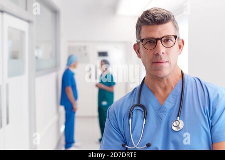 Portrait Von Reife Männliche Arzt Tragen Scrubs Stehen In Beschäftigt Krankenhauskorridor Stockfoto