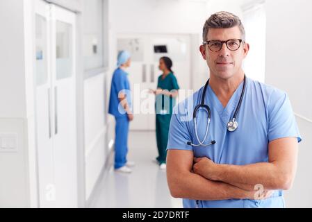 Portrait Von Reife Männliche Arzt Tragen Scrubs Stehen In Beschäftigt Krankenhauskorridor Stockfoto