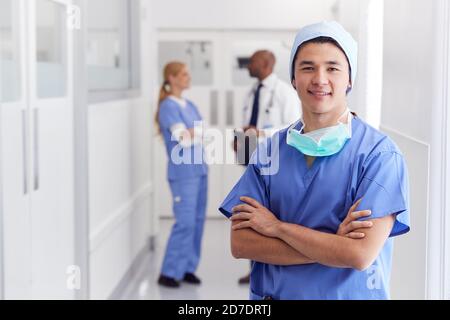 Porträt Des Männlichen Arztes Trägt Scrubs Stehen In Busy Hospital Korridor Stockfoto