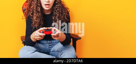 Frau, die hinter einem Spielstuhl in ihren Händen sitzt und rot hält Gamepad auf gelbem Hintergrund Stockfoto