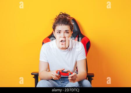 Frau, die hinter einem Spielstuhl in ihren Händen sitzt und rot hält Gamepad auf gelbem Hintergrund Stockfoto
