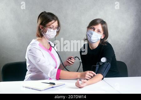 Eine junge Ärztin in einem medizinischen Gewand und Maske Misst den Blutdruck mit einem Tonometer an eine maskierte Frau Stockfoto