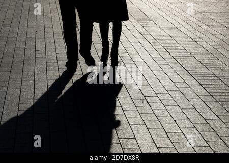 Ein Paar, das die Straße entlang geht, Silhouetten und Schatten von Mann und Frau auf dem Fußgängerweg. Männliche und weibliche Beine, Beziehungskonzept Stockfoto