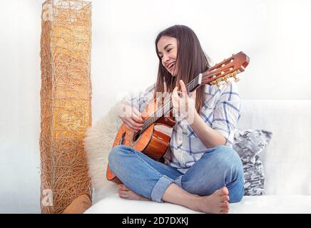 Eine junge Frau im Wohnzimmer mit Gitarre sitzt auf einem leichten Sofa und spielt. Stockfoto