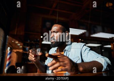 Gutaussehender afroamerikanischer Mann, der Zigarre raucht und Whisky trinkt Stockfoto