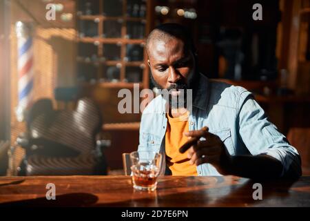 Ein gutaussehender afroamerikanischer Mann raucht Zigarre an der Bar des Barbershops Stockfoto