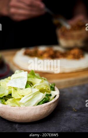 Nahaufnahme einer Schüssel mit gehacktem Salat auf einem Tisch, und ein junger Mann im Hintergrund bereitet ein Durum oder Burrito, mit Hühnerfleisch gekocht mit Dif Stockfoto