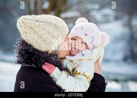 Die junge Mutter umarmt ihre kleine Tochter und küsst sie auf die Wange. Winterzeit, Outdoor-Aktivitäten Stockfoto