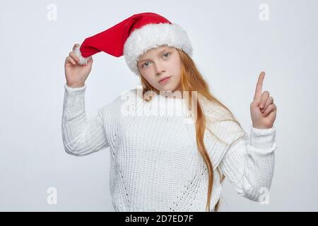 Attraktive Teenager-Mädchen mit roten Haaren trägt einen Weihnachtsmann Hut. Zeigt mit dem Finger eine leere Stelle für Text. Foto-Sitzung im Studio auf einem weißen Stockfoto