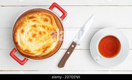 Hausgemachter Hüttenkäse Käsekuchen in roter Keramikschale und Tasse Tee mit Zitrone auf weißem Holztisch. Draufsicht. Stockfoto