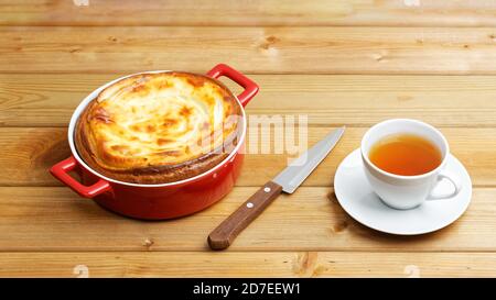 Hausgemachter Hüttenkäse Käsekuchen in roter Keramikschale und Tasse Von Tee auf Holztisch Stockfoto