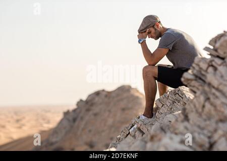 Depression und Einsamkeit. Trauriger junger Mann, der allein auf einer Bergklippe sitzt und die Wüste überblickt. Stockfoto