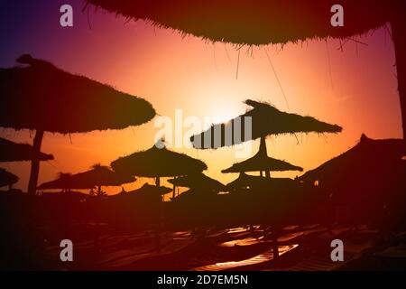 Wunderschönes Sonnenlicht bei Sonnenuntergang am Strand. Abend im Urlaub am Meer, Kopierplatz. Die untergehende Sonne beleuchtet den Strand Silhouette Sonnenschirme, w Stockfoto