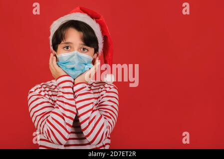 Frohe Weihnachten, lustiges Kind mit medizinischer Maske trägt Weihnachtsmann Hut Auf rotem Hintergrund Stockfoto