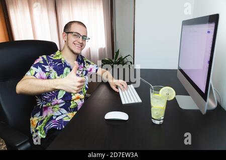 Junger Mann, der am Computer arbeitet, mit hawaiiihemd Fernarbeit, mit einem erfrischenden Getränk. Entspannender Job. Urlaub und Arbeit kombiniert. Lächelnd an Stockfoto