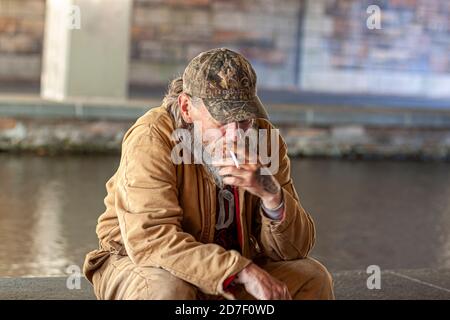 Frederick, MD, USA 10/14/2020: Ein älterer Kaukasusmann mit Baseballmütze und brauner Jacke sitzt an einem Fluss unter einer Brücke und raucht eine Zigarette. Stockfoto