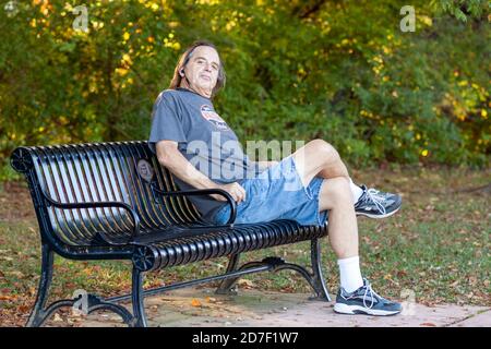 Frederick, MD, USA 10/14/2020: Langhaariger Mann mittleren Alters entspannt sich nach einer Wanderung auf einer Parkbank. Er dreht seinen Kopf, während er mit gekreuzten Beinen sitzt Stockfoto