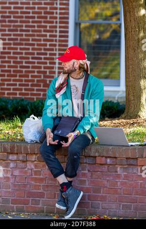 Frederick, MD, USA 10/14/2020: Ein freimütiger junger blonder Mann mit Bandana unter seinem Baseballhut sitzt auf einer Wand auf der Straße. Er hat sein l Stockfoto