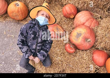 Junge im Halloween Kostüm mit Kürbissen auf dem Bauernmarkt steht auf Heu. Furchterregende Dekorationen. Kinder Trick or Treat. Das Tragen einer schützenden Gesichtsmaske an Halloween Stockfoto