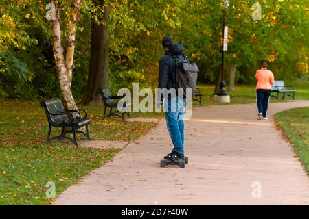 Der junge Mann mit Hoodie, Rucksack und Hut fährt im Baker Park auf einem Betonweg Skateboarding. Das Bild wurde im Herbst mit gefallenen Blättern aufgenommen Stockfoto