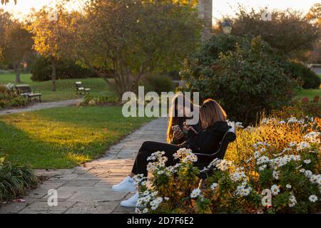 Frederick, MD, USA 10/14/2020: Zwei Teenager-Mädchen sitzen bei Sonnenuntergang auf einer Bank im Baker Park. Beide betrachten ihre Smartphones. Da ist ar Stockfoto