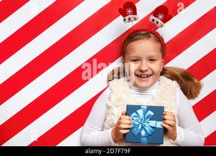 Glückliches kleines Mädchen hält ein Weihnachtsgeschenk in ihren Händen. Kind in Fäustlingen Stirnband auf einem rot-weißen Hintergrund. Stockfoto