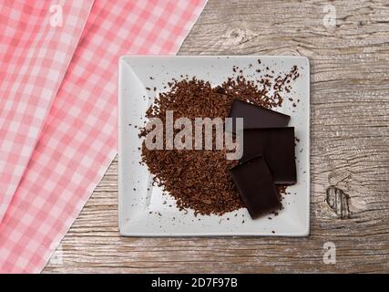 Geriebene Schokolade auf einem weißen Teller. Teller steht auf altem Holz und karierter Tischdecke. Dunkle Schokolade von oben fotografiert. Stockfoto