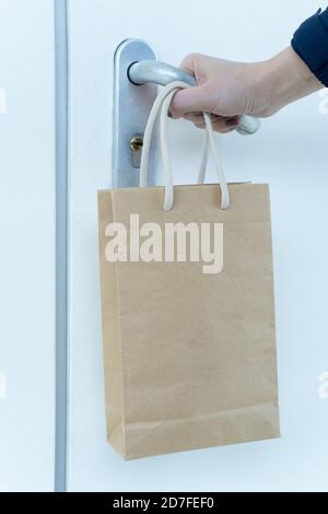 Eine menschliche Hand versucht, eine verschlossene Tür zu öffnen und eine Papiertüte mit Lebensmitteln hängt an seinem Handgelenk. Kontaktlose Lebensmittelzustellung. Online-Bestellung von Produkten Stockfoto