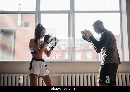 Ein Mann mit Boxpfoten an den Händen lehrt die Technik des Schlagens ein Anfänger Mädchen in einem leichten Fitnessstudio Stockfoto