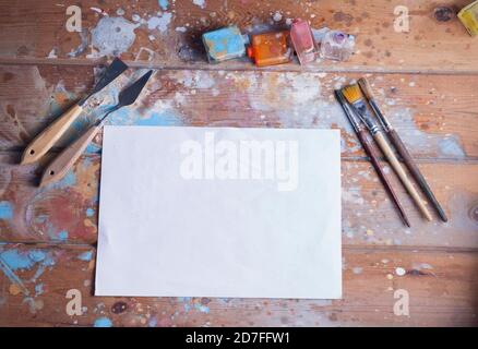 Künstlerschreibtisch und Materialmodell. Verschiedene Pinsel und Palettenmesser. Das Bild enthält ein leeres weißes Blatt Papier, ideal für ein kreatives Modell. Stockfoto