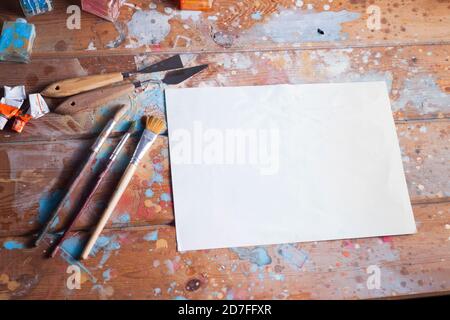 Künstlerschreibtisch und Materialmodell. Verschiedene Pinsel und Palettenmesser. Das Bild enthält ein leeres weißes Blatt Papier, ideal für ein kreatives Modell. Stockfoto