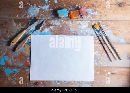 Künstlerschreibtisch und Materialmodell. Verschiedene Pinsel und Palettenmesser. Das Bild enthält ein leeres weißes Blatt Papier, ideal für ein kreatives Modell. Stockfoto