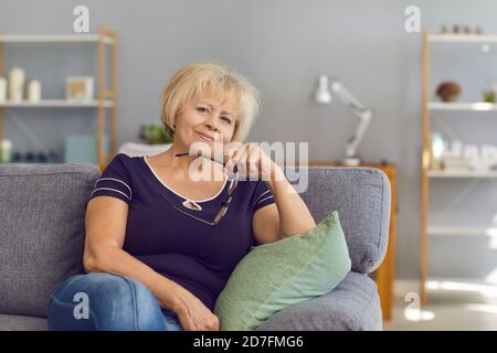 Reife Frau lächelt und schaut auf die Kamera, während sie auf einem Sofa im Wohnzimmer sitzt. Stockfoto