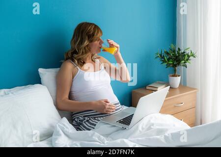 Eine lächelnde und glückliche schwangere Kaukasierin in sitzender Position in ihrem Bett, während sie an ihrem Laptop arbeitet. Eine Hand liegt auf ihrem Bauch und sie trinkt Stockfoto