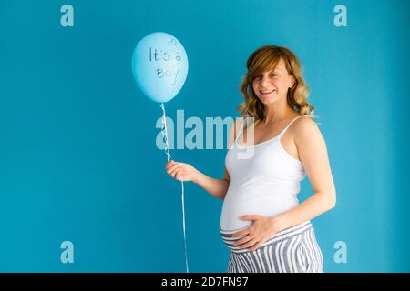 Eine schwangere lächelnde Frau hält einen Ballon mit den Worten "es ist ein Junge". Sie ist vor einer blauen Wand. Stockfoto