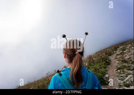 Mädchen von hinten mit Pflanzenhörnern auf dem Kopf in einer blauen Jacke im Nebel in den Bergen. Fun Konzept, Wandern in den Bergen, Trekking, aktiv l Stockfoto